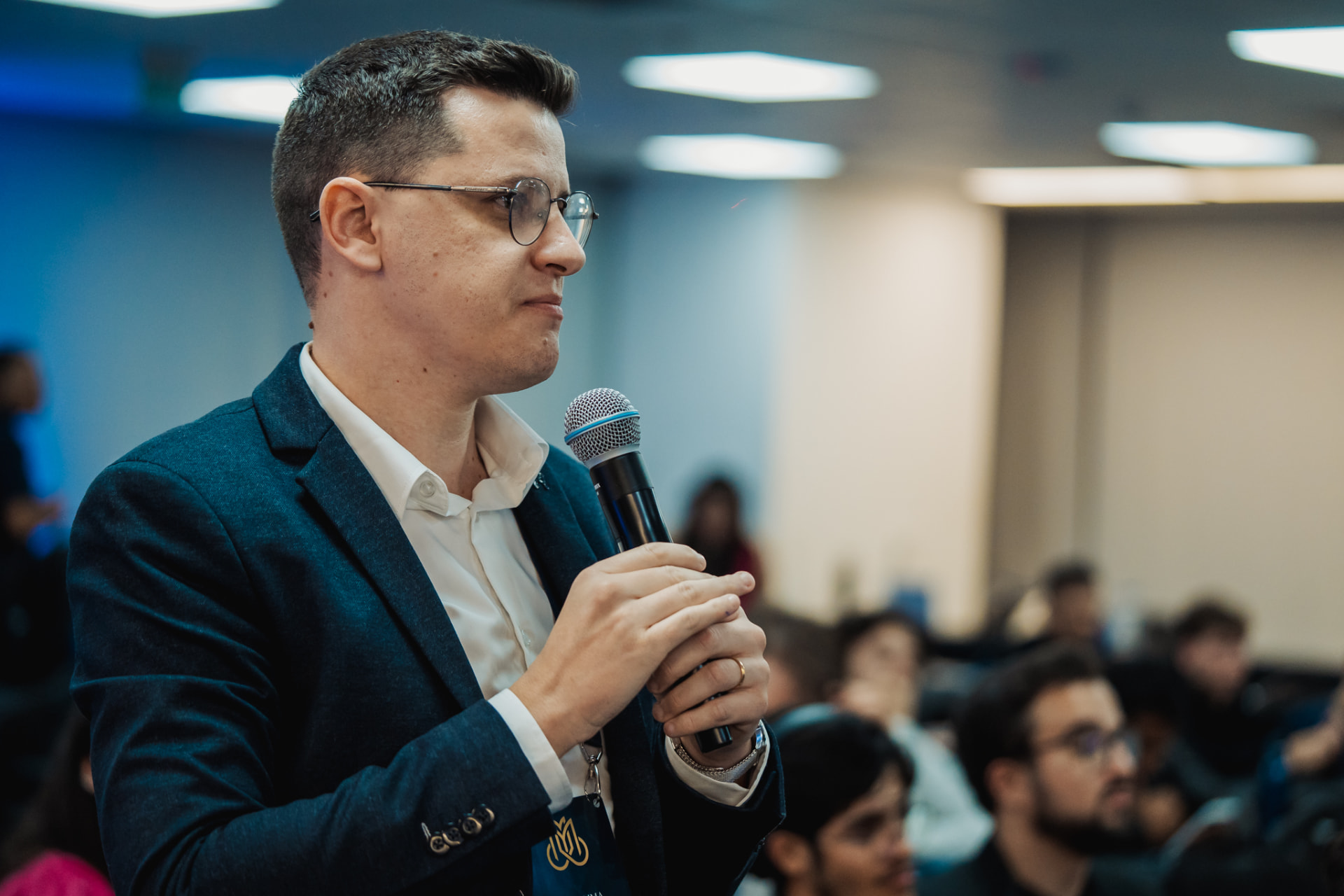 João de blazer e óculos, segurando um microfone durante uma palestra